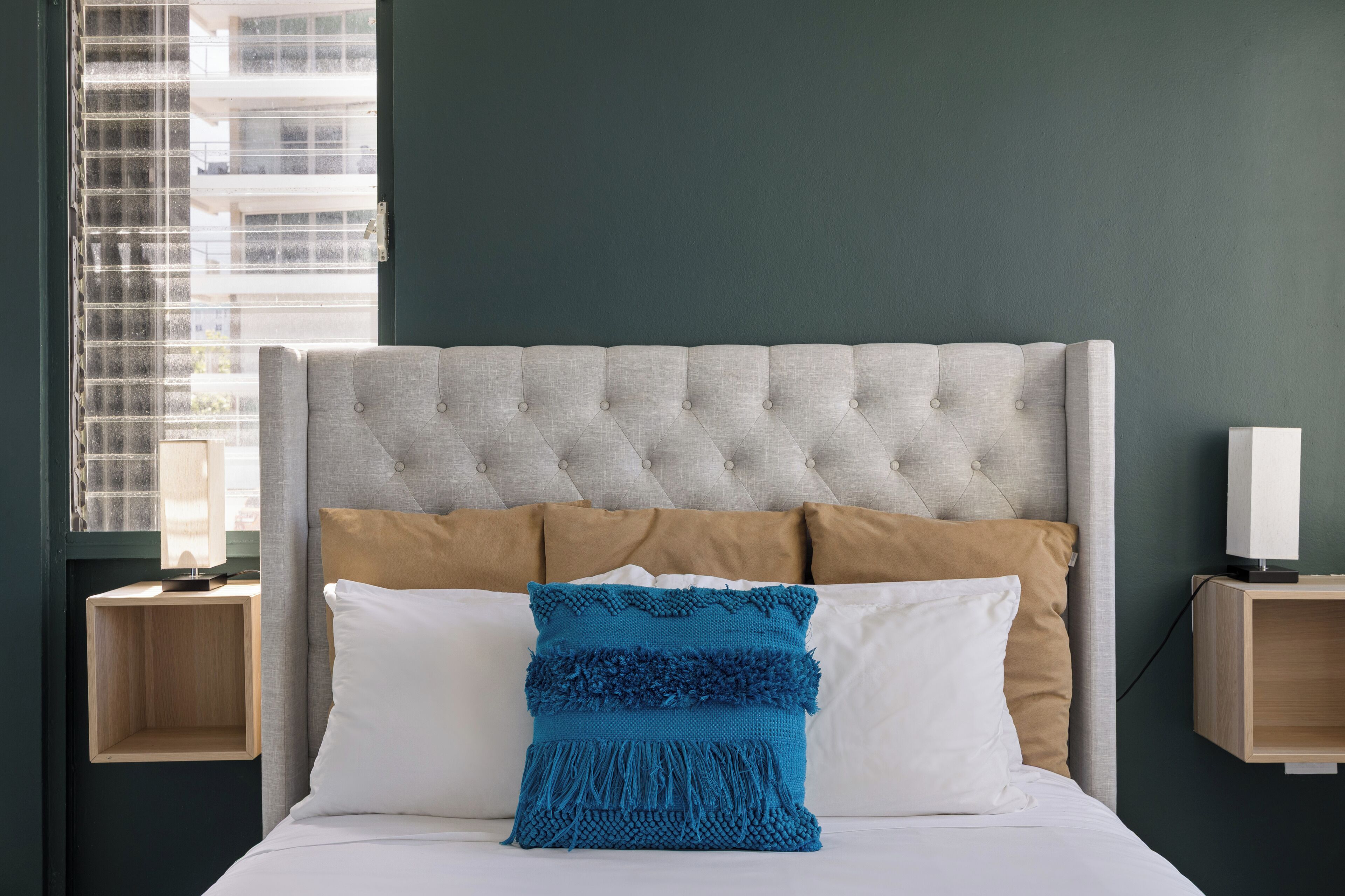 A neatly made bed with a tufted headboard, two pillows, and a blue decorative pillow. There are two bedside tables with a lamp on one.
