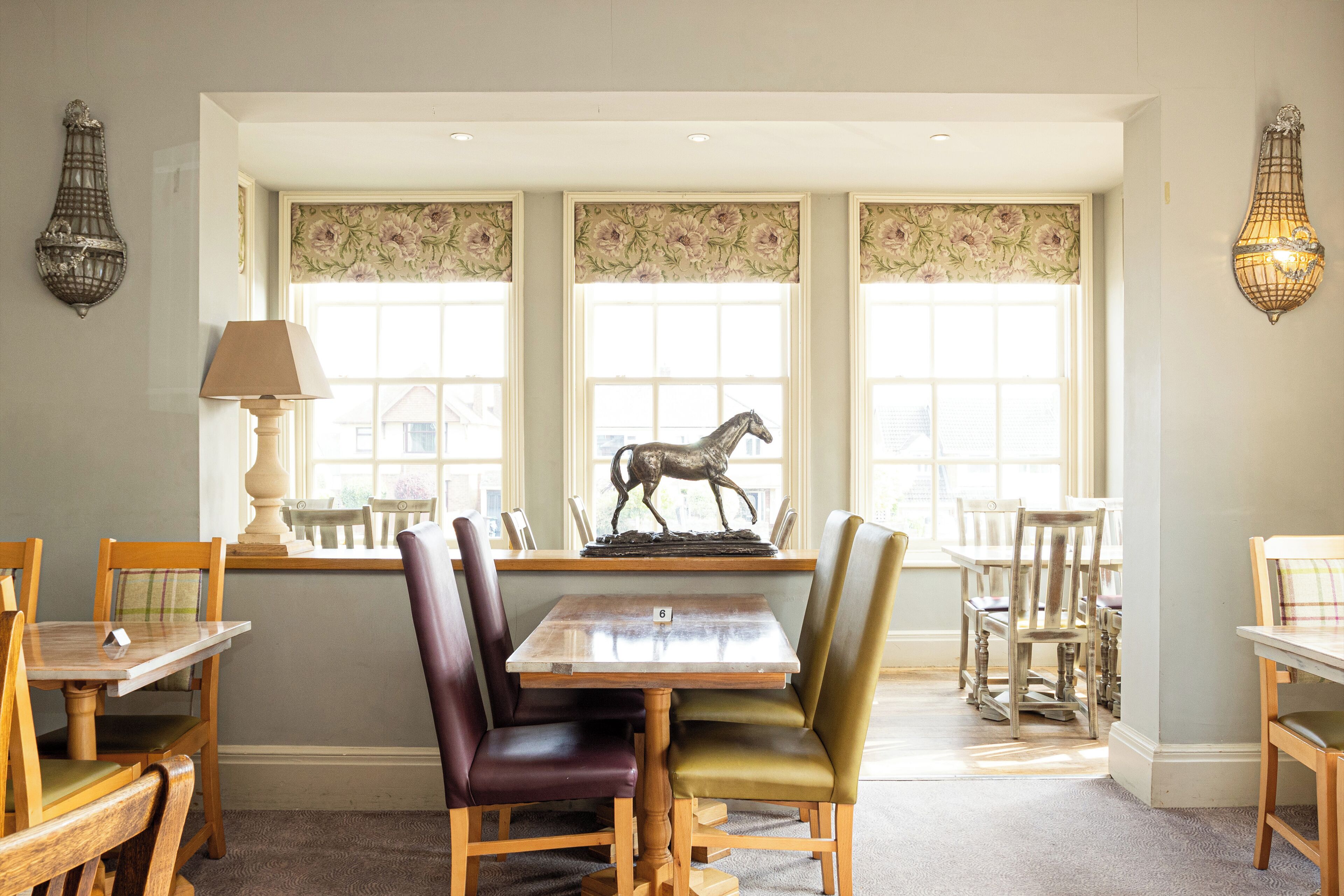 Un comedor con mesa y sillas de madera, un centro de mesa con escultura de caballo y cortinas de encaje en las ventanas.