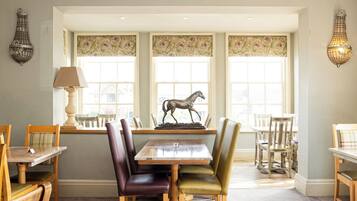 Un comedor con mesa y sillas de madera, un centro de mesa con escultura de caballo y cortinas de encaje en las ventanas.