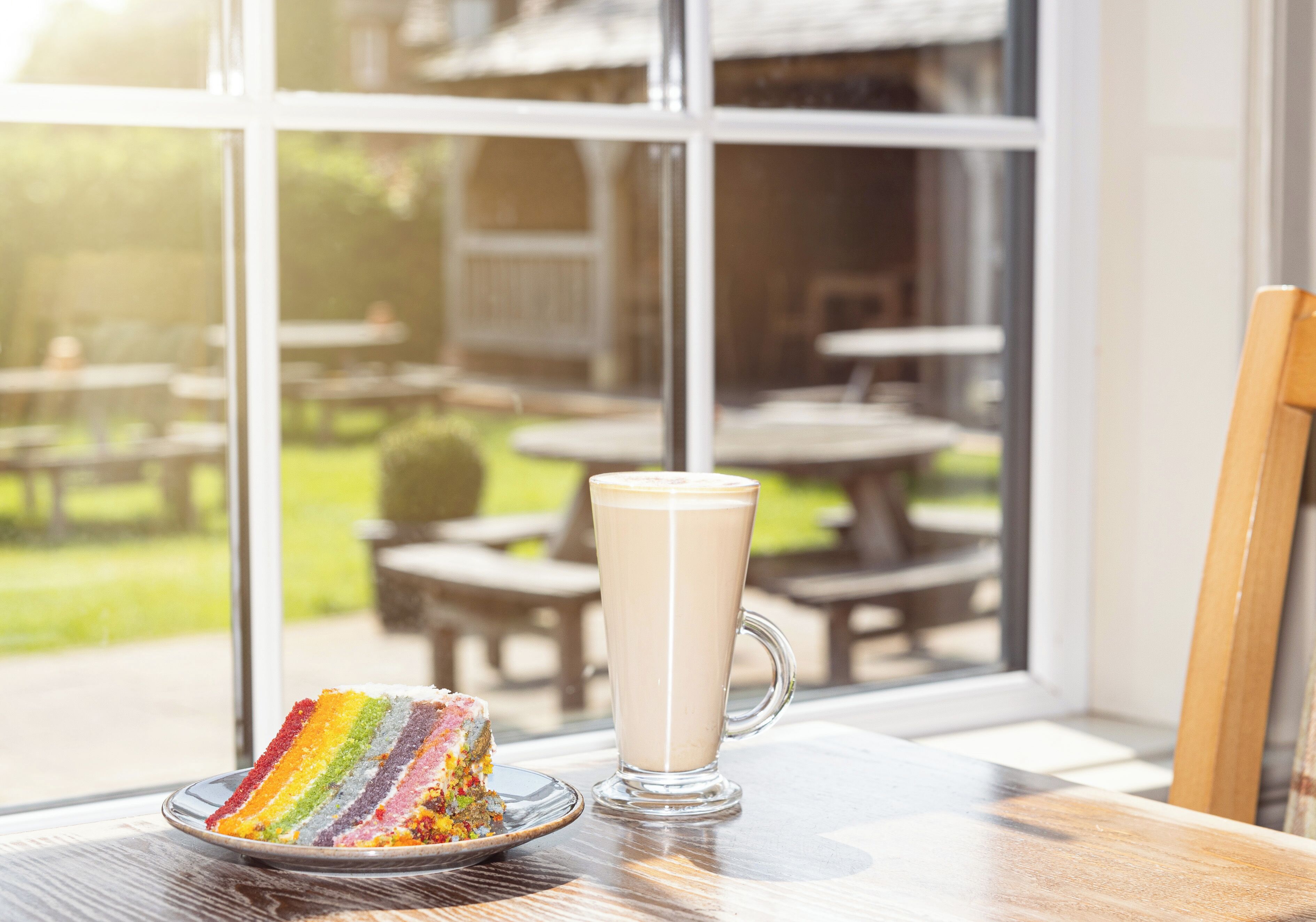 Une part de gâteau arc-en-ciel sur une assiette et un verre de lait sur une table près d’une fenêtre.