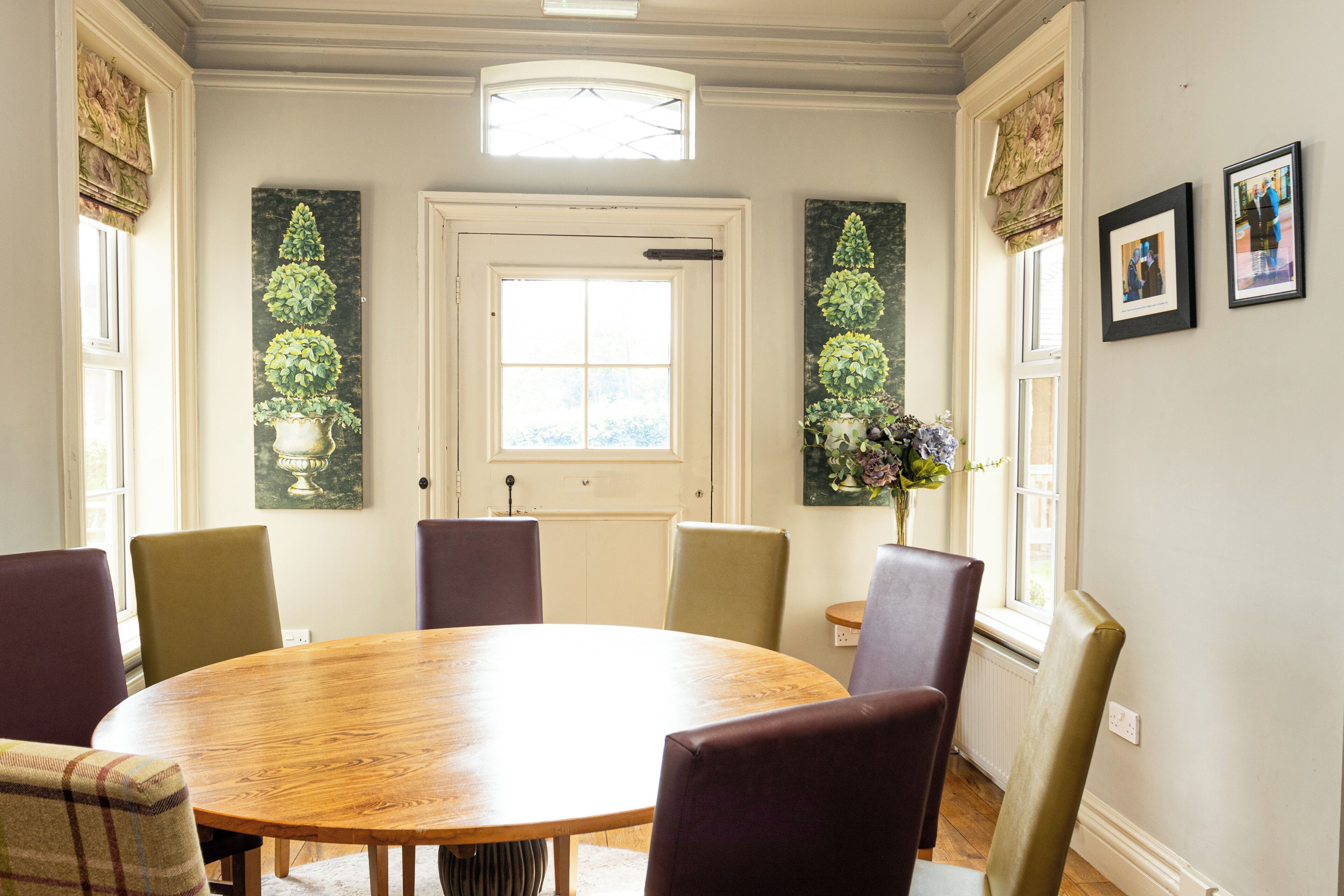 A dining room with a round wooden table, six chairs, and two windows with curtains.