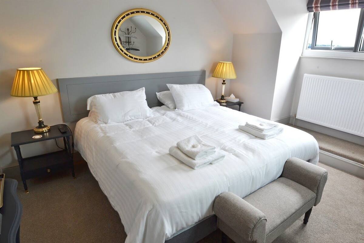 A neatly made bed with white linens, two bedside lamps, a round mirror, and a bench at the foot of the bed.