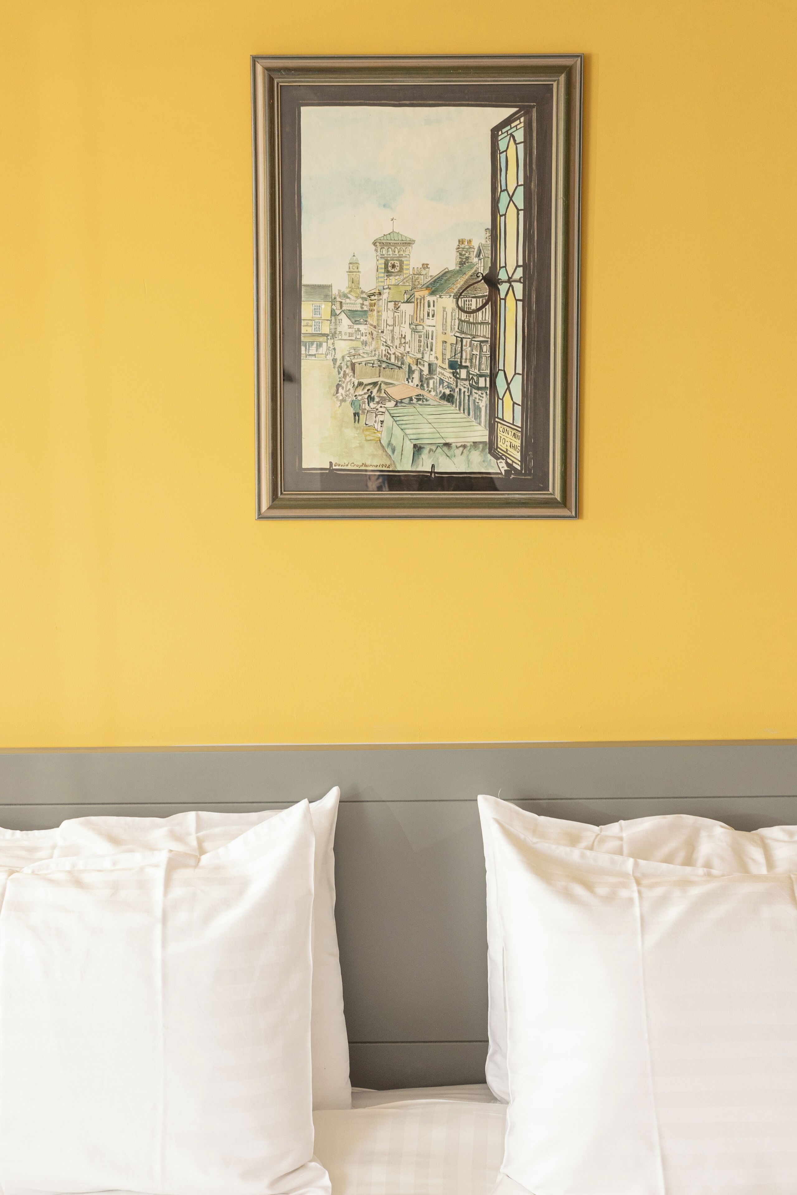 A framed artwork of a street scene on a yellow wall above a bed with white pillows and a gray headboard.