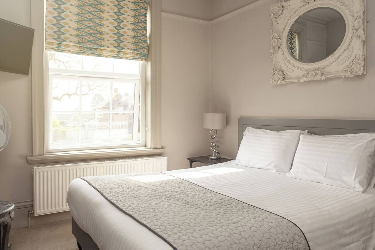 A neatly arranged bedroom with a large bed, a bedside table with a lamp, a window with patterned curtains, and a mirror on the wall.