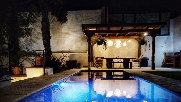 Una piscina con pérgola y zona de estar, rodeada de plantas en macetas y un muro de piedra.