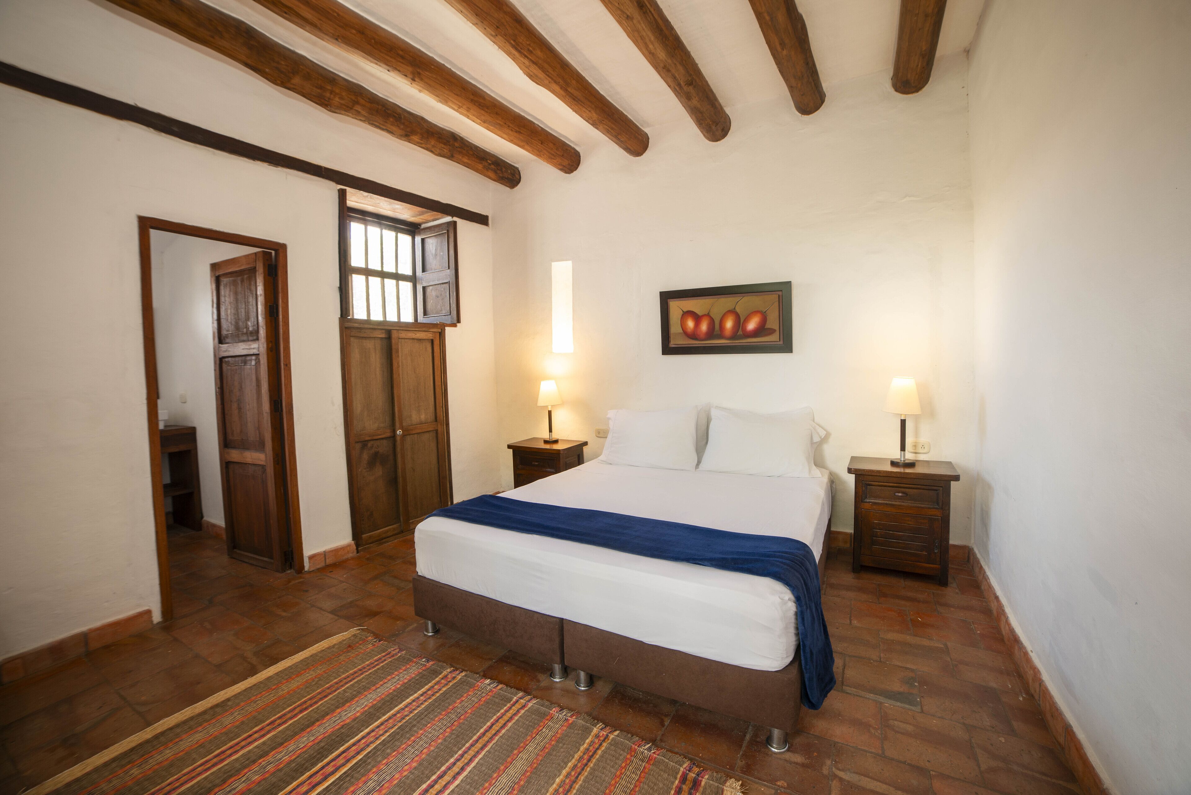 A bedroom with a bed, wooden beams, a door, a wall-mounted picture, and two bedside tables with lamps.