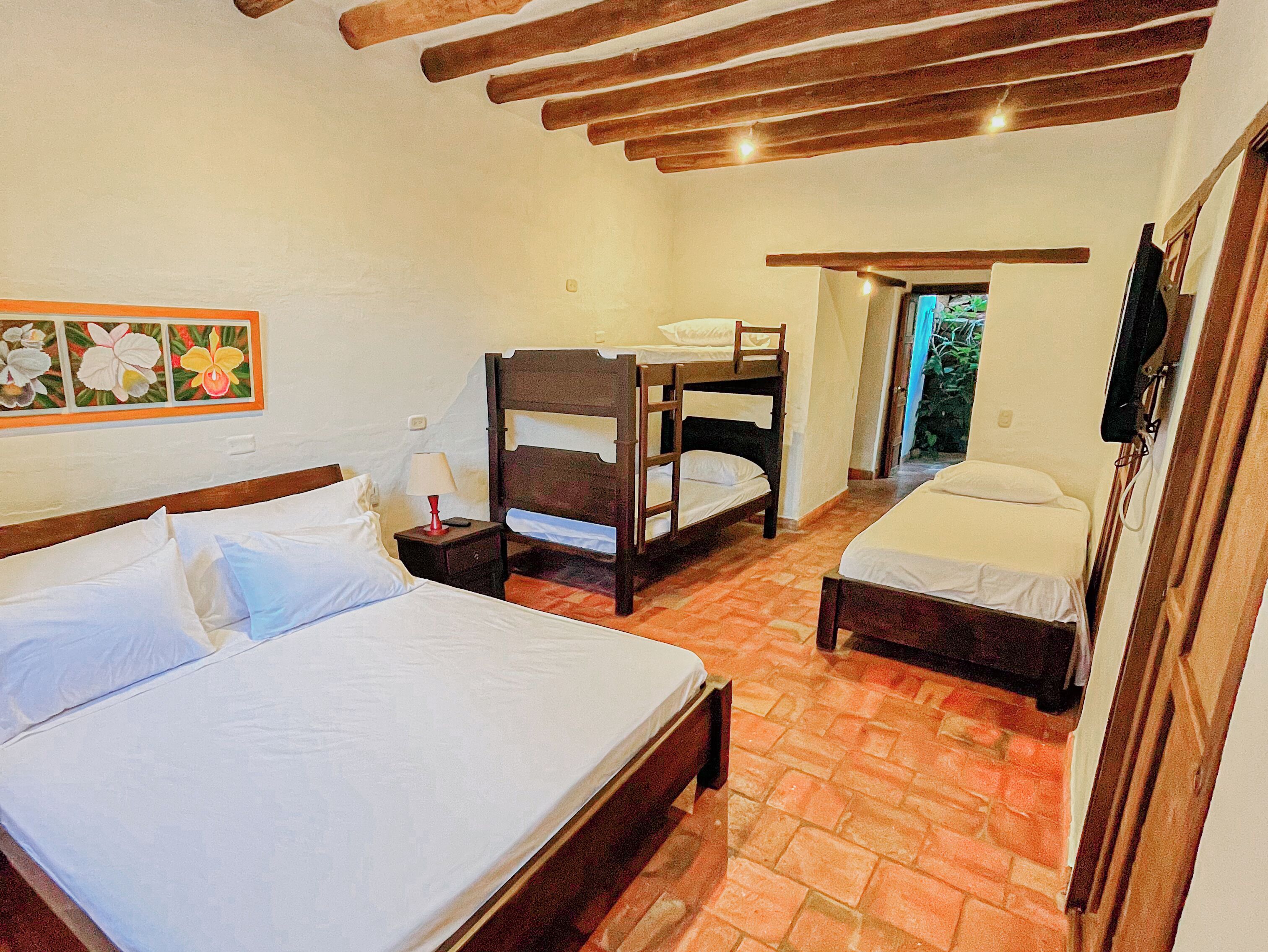 Un dormitorio con una cama grande, dos camas más pequeñas, una mesita de noche con lámpara y techo de madera.