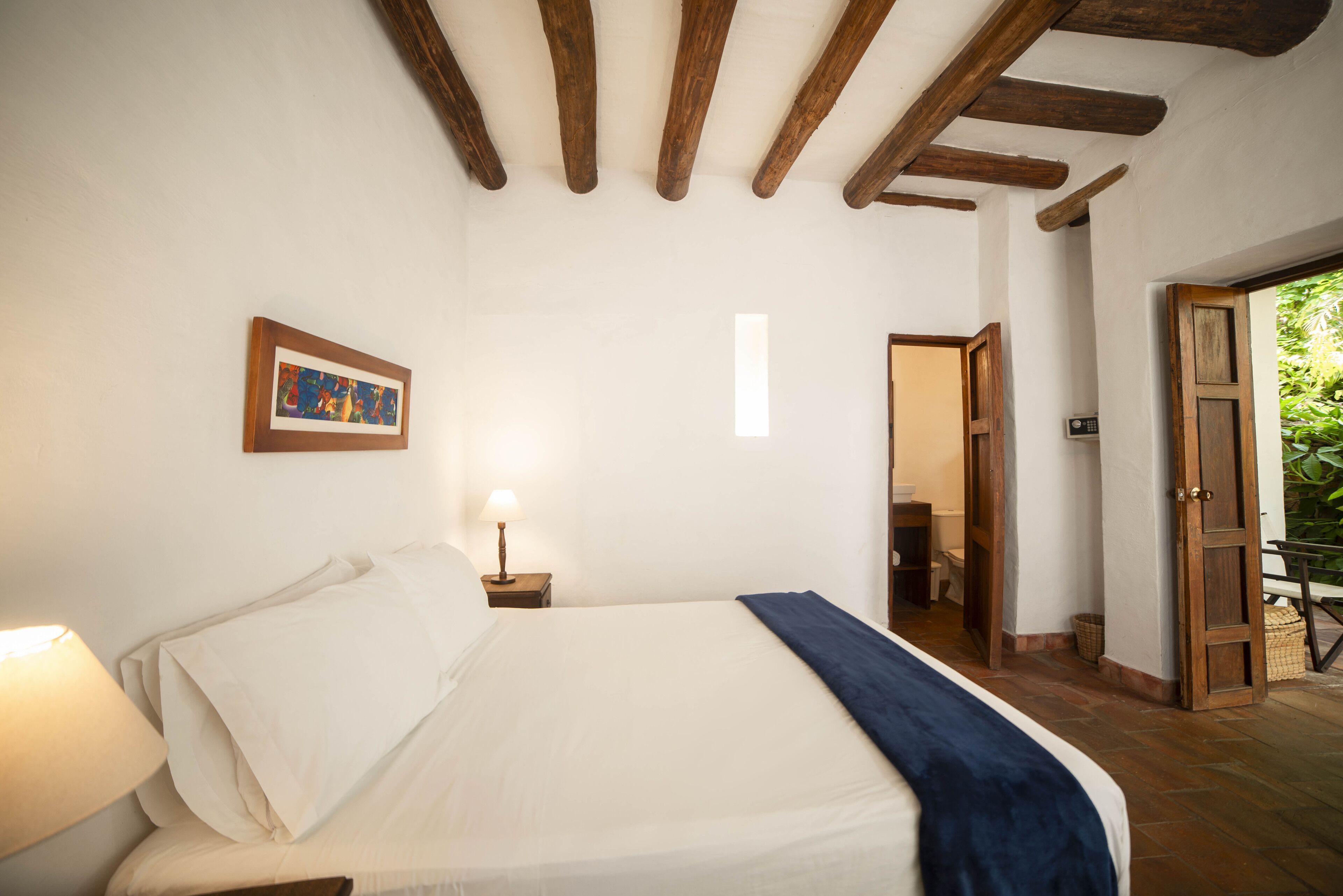 A bedroom with a bed, a nightstand with a lamp, a framed picture on the wall, and a wooden door leading to another room.