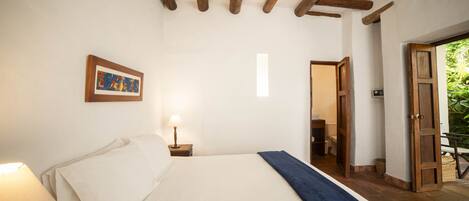 A bedroom with a bed, a nightstand with a lamp, a framed picture on the wall, and a wooden door leading to another room.