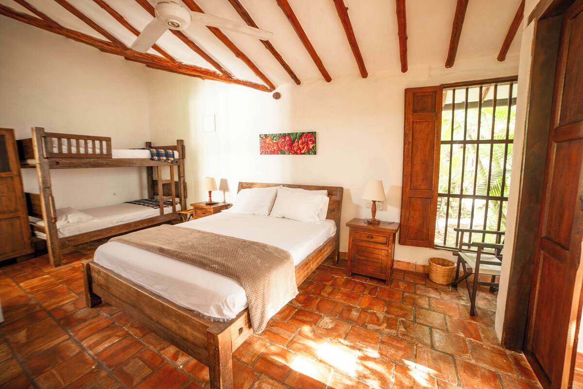 A bedroom with a wooden bed, a lower bunk bed, a nightstand, a lamp, a wooden door, and a window with bars.