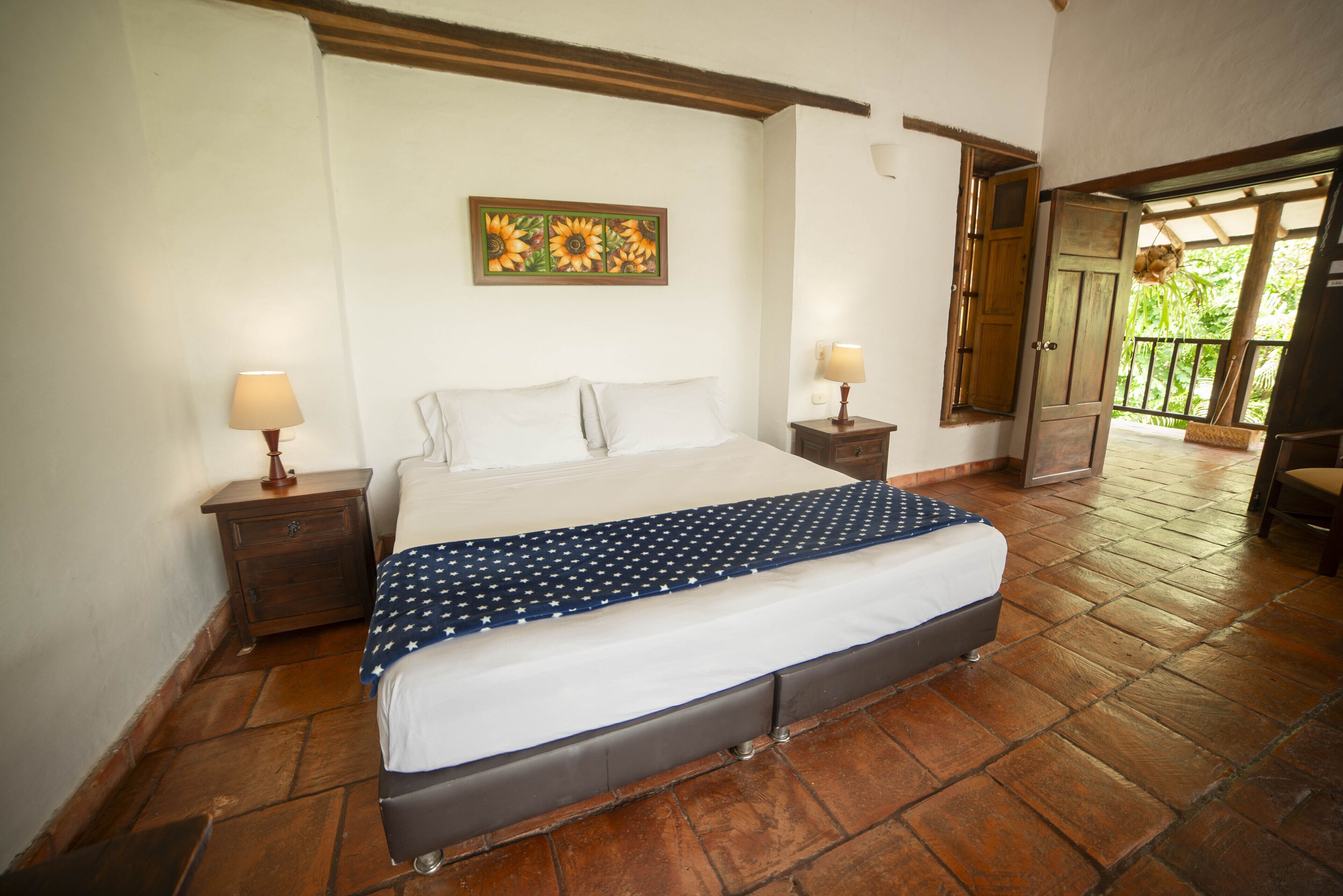 Een slaapkamer met een groot bed, twee nachtkastjes, een schilderij aan de muur, en een houten deur die leidt naar een buitenruimte.