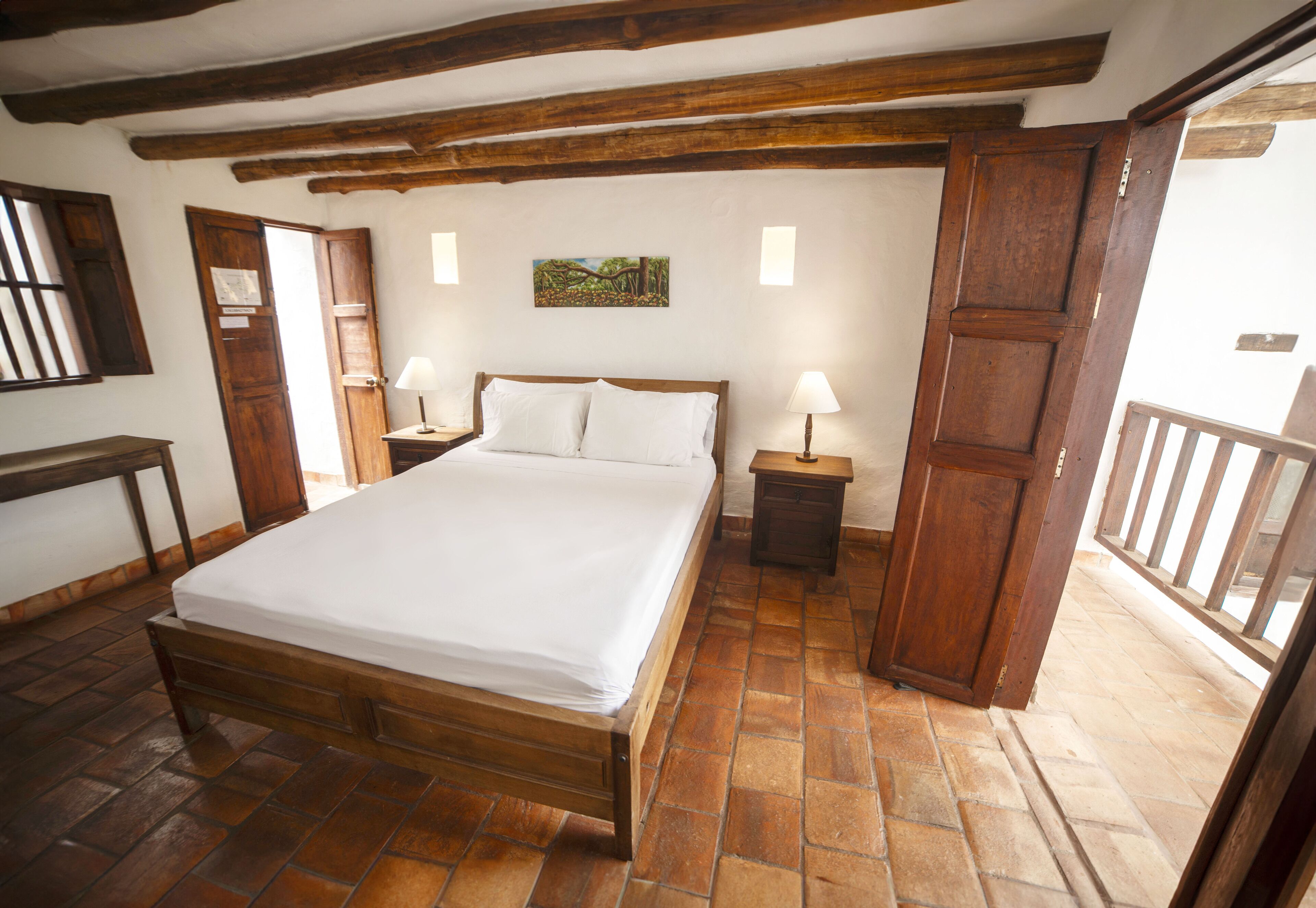 A bedroom with a wooden bed, two bedside tables with lamps, a wooden door, and a balcony with a railing.