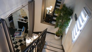 Eine Treppe mit schwarz-weiß kariertem Boden, ein weißer Stuhl mit farbigem Muster, ein Topfpflanze und ein Spiegel, der eine Chandeliebre widergibt.