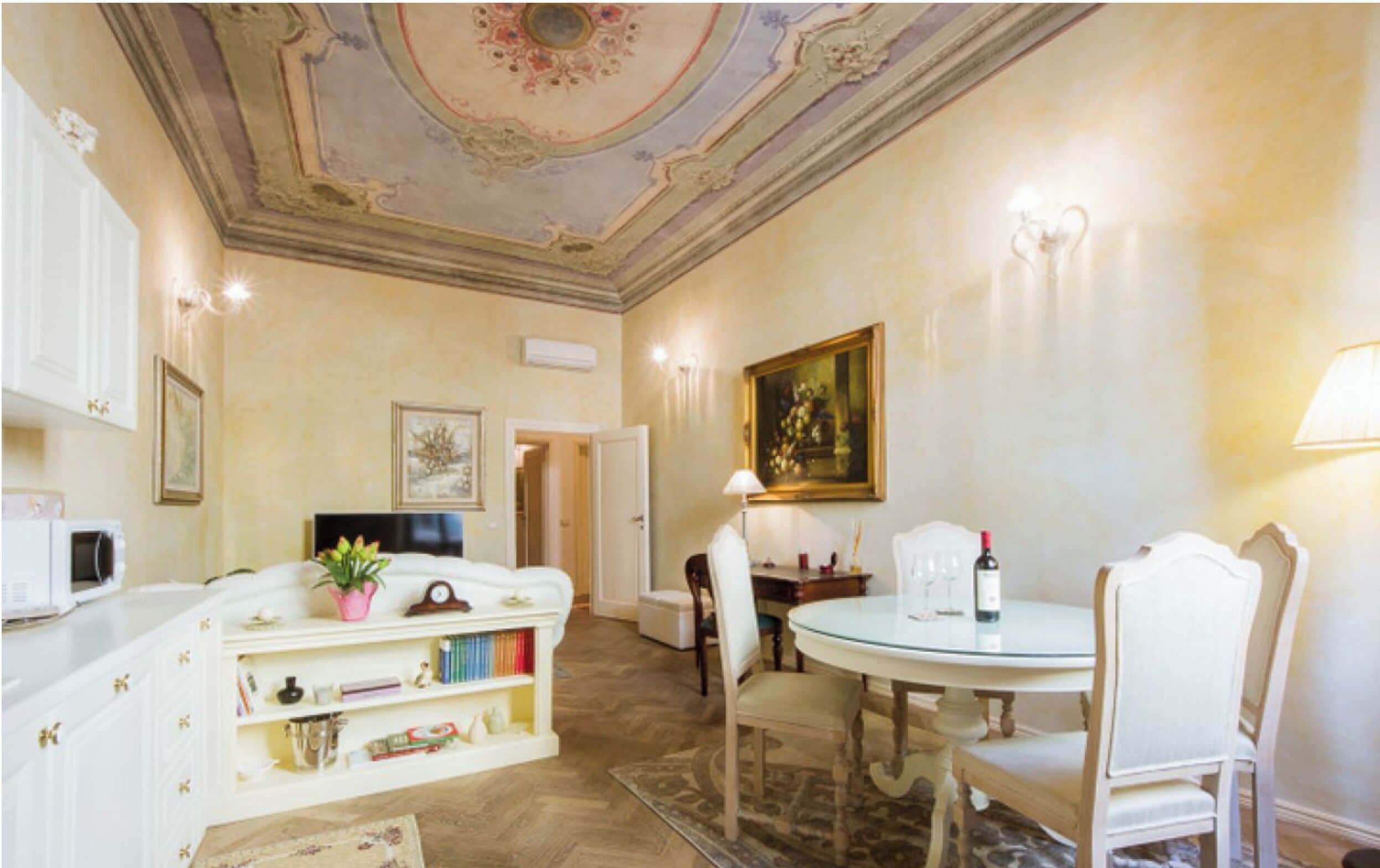 Une pièce avec un canapé blanc, une bibliothèque, une table à manger avec des chaises et un plafond orné de moulures décoratives.