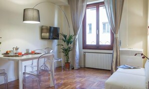 A room with a dining table, chairs, a TV, a potted plant, a radiator, and a window with curtains. - Apartments Florence- Santa Maria Suite (Firenze)
