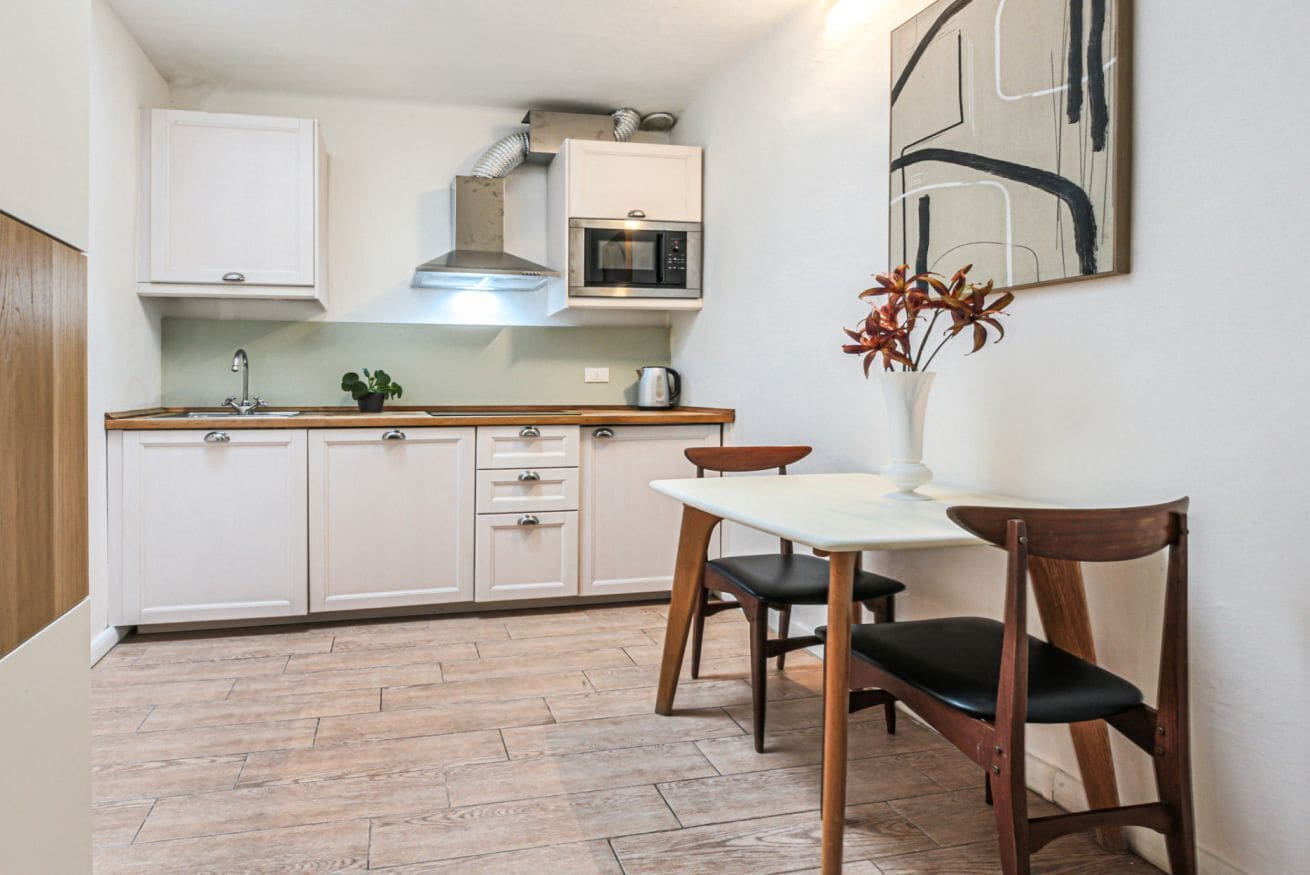 A compact kitchen with white cabinets, a microwave, and a stove hood. There is a dining table with two chairs and a vase with flowers on the wall.
