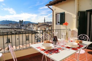 Une table de balcon, dressée avec de la vaisselle, des verres et une bouteille, d’où l’on peut admirer un paysage urbain avec ses bâtiments et son monument.