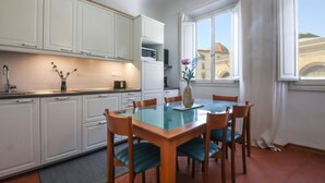 Une cuisine compacte avec des meubles blancs, une table à manger avec des chaises et une fenêtre donnant sur des immeubles.