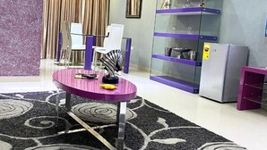Um quarto com uma mesa roxa, cadeiras, um armário de vidro e uma geladeira.