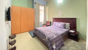 Quarto com cama, mesa de cabeceira, cadeira, guarda-roupa, ventoinha no teto e janela com cortinas.