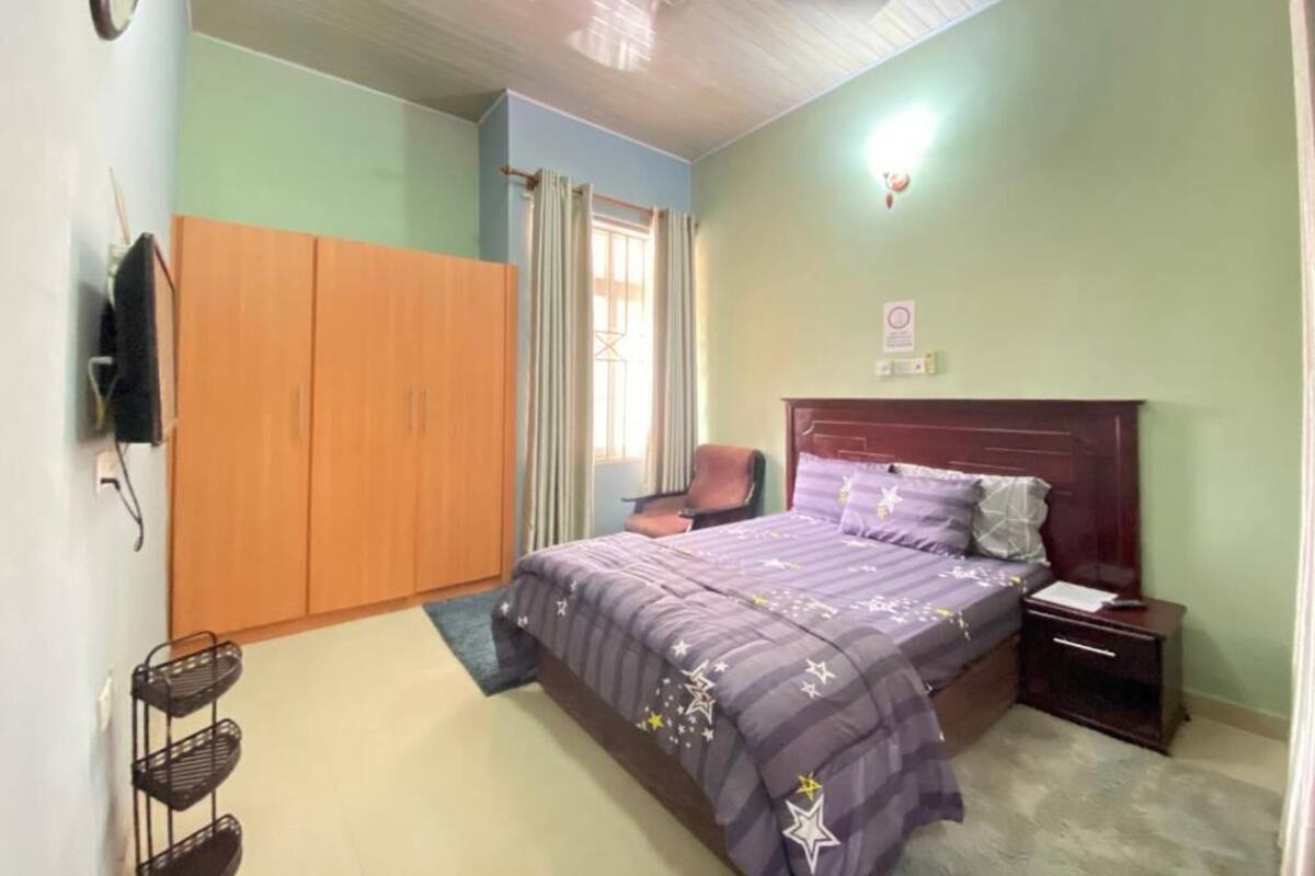 Ein Schlafzimmer mit Bett, Nachttisch, Stuhl, Kleiderschrank, Deckenventilator und einem Fenster mit Vorhängen.