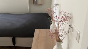 Una stanza piccola con un letto, un comodino con un vaso e tre mensole a parete con barattoli.
