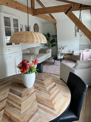 Un salón acogedor con una mesa redonda de madera, un armario blanco, un sofá y una lámpara colgante de mimbre.