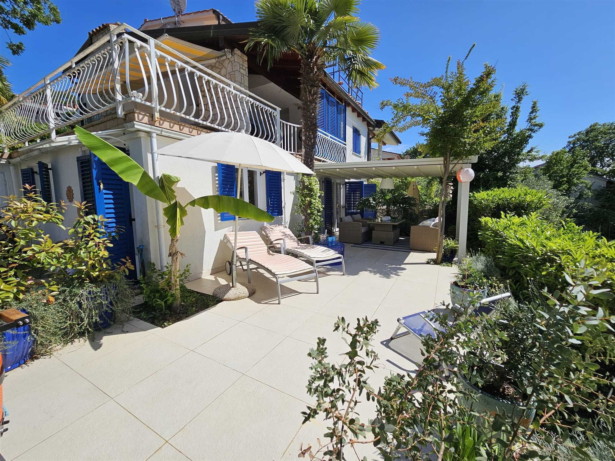 A two-story house with a balcony, a covered patio area with outdoor furniture, and lush greenery.