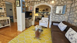 A living room with a stone wall, a sofa, a TV, and a kitchen visible through an archway. - Apartment Barbara with sea view (Njivice)