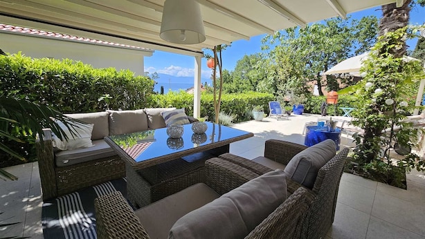 A covered outdoor seating area with wicker furniture and a glass table, surrounded by greenery and a view of the sea. - Apartment Barbara with sea view (Njivice)