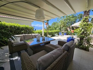 A covered outdoor seating area with wicker furniture and a glass table, surrounded by greenery and a view of the sea. - Apartment Barbara with sea view (Njivice)