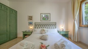 A neatly made bed with towels and a floral arrangement, a green wardrobe, and a framed picture on the wall.