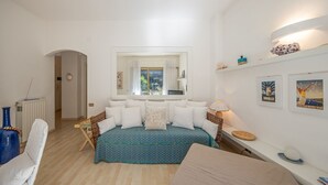 A bedroom with a bed, a chair, a small table, a shelf with decorative items, and a window with curtains.