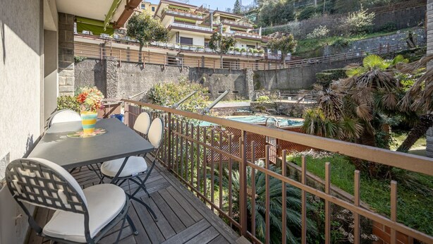A balcony with a table and chairs, overlooking a garden and a hillside with buildings.