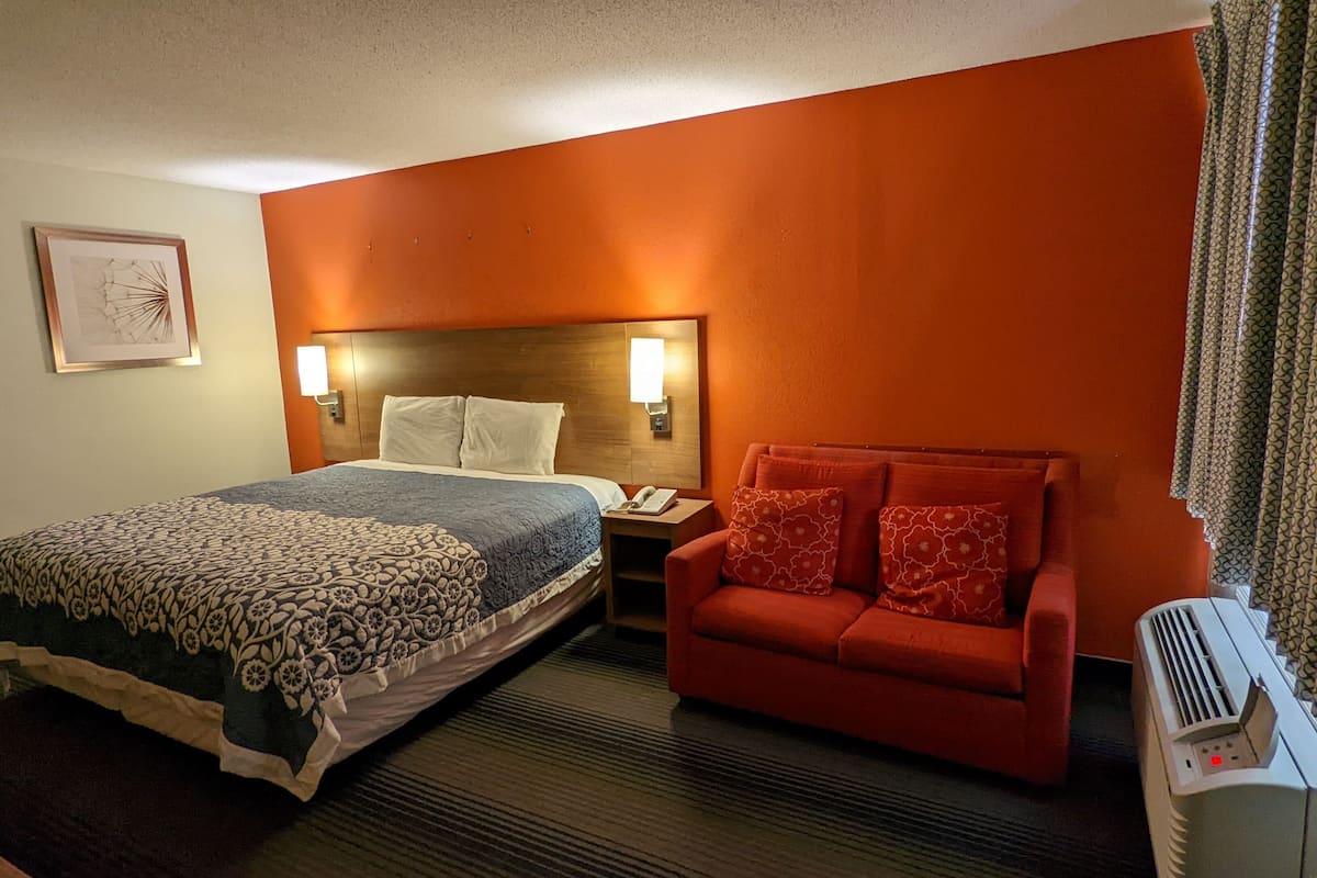 Una habitación de hotel con una cama, un sofá rojo, una mesita de noche, una lámpara y un aire acondicionado instalado en la pared.