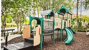 Un parque infantil con estructuras verdes y beige, que incluyen un tobogán y zonas de escalada, rodeado de árboles y mantillo.
