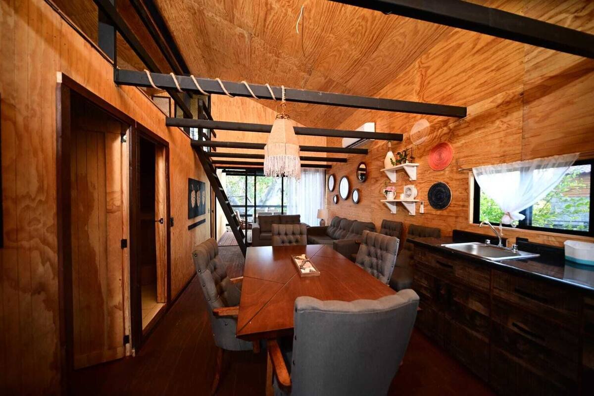 Una cocina moderna con detalles de madera, un comedor con una mesa grande y varias sillas, y vistas al exterior con vegetación.
