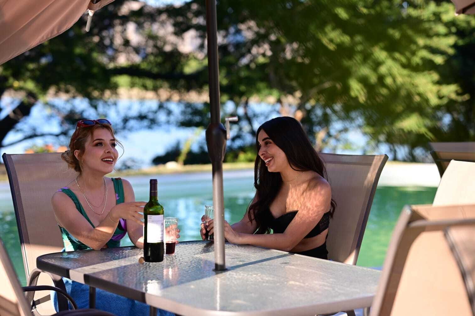 Due persone sedute a un tavolo all'aperto con una bottiglia di vino e bicchieri, con vista su un corpo d'acqua.