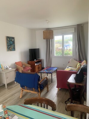 En stue med et fjernsyn, fletningsmøbler, et bord med en farverig bordløber, og et vindue med gardiner.