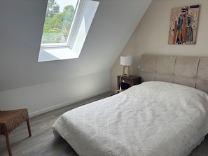A bedroom with a skylight, a bed with a patterned bedspread, a nightstand with a lamp, and a painting on the wall.