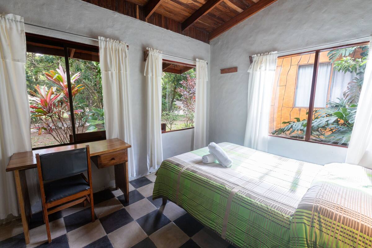 A room with a bed, a desk, and a chair. There are windows with curtains, and a view of greenery outside.