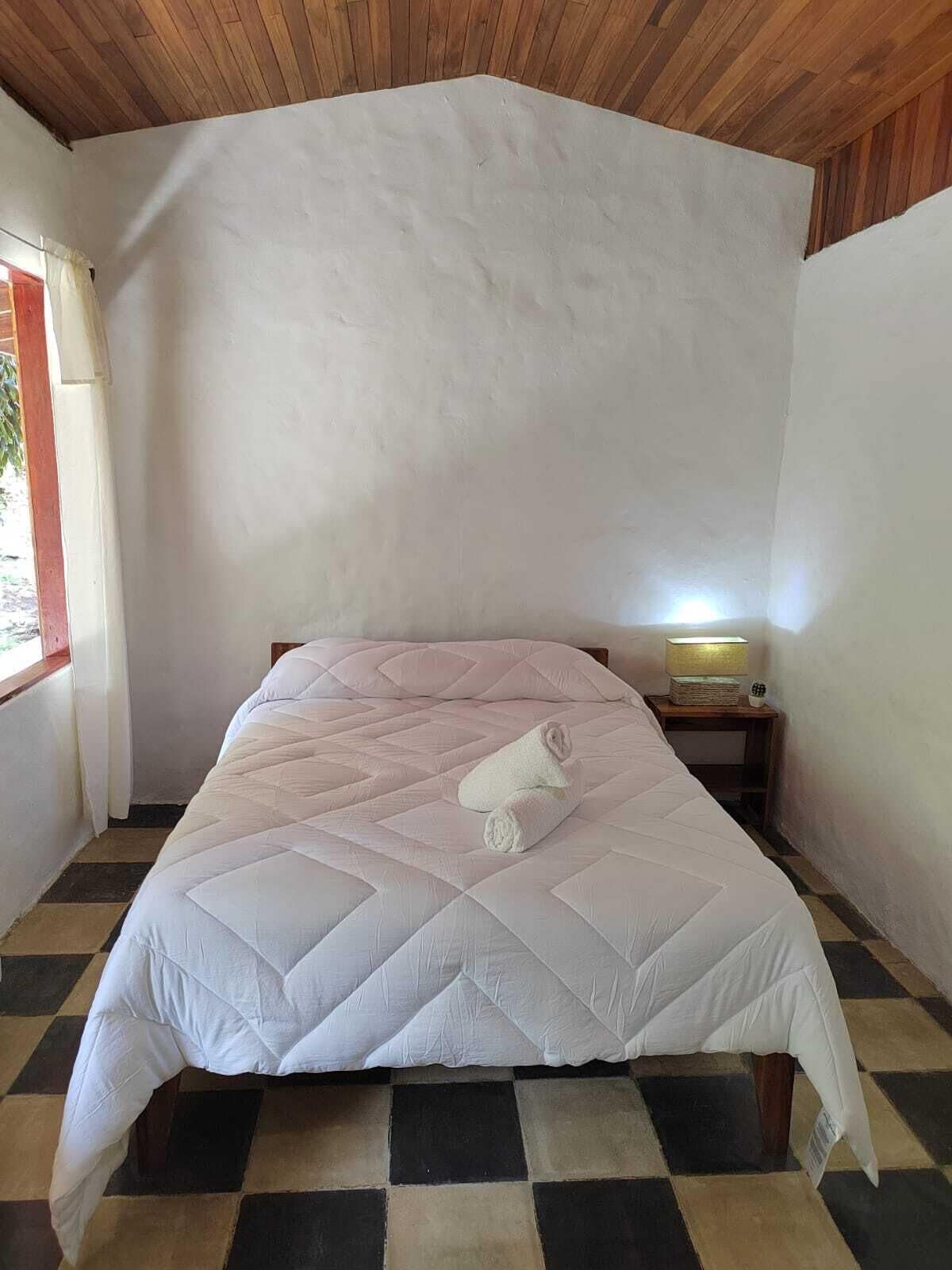 A single bed with a quilted white bedspread, a rolled towel, and a bedside lamp on a wooden nightstand. The room has a checkered floor and a wooden ceiling.
