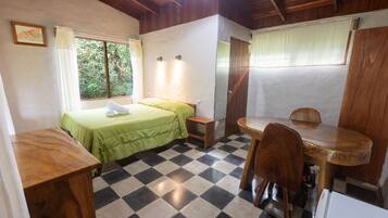 A room with a bed, a small table, and a chair. There is a window with curtains, a door, and a checkered floor.