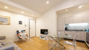 A modern living room with a glass table and chairs, a grey sofa, a TV, and a kitchen area with cabinets and a sink. - Charming View apartment by Mmega (Firenze)