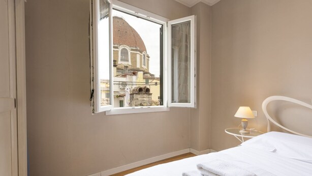 A hotel room with a bed, a window with a view of a building, a bedside table with a lamp, and a wall-mounted radiator. - Charming View apartment by Mmega (Firenze)