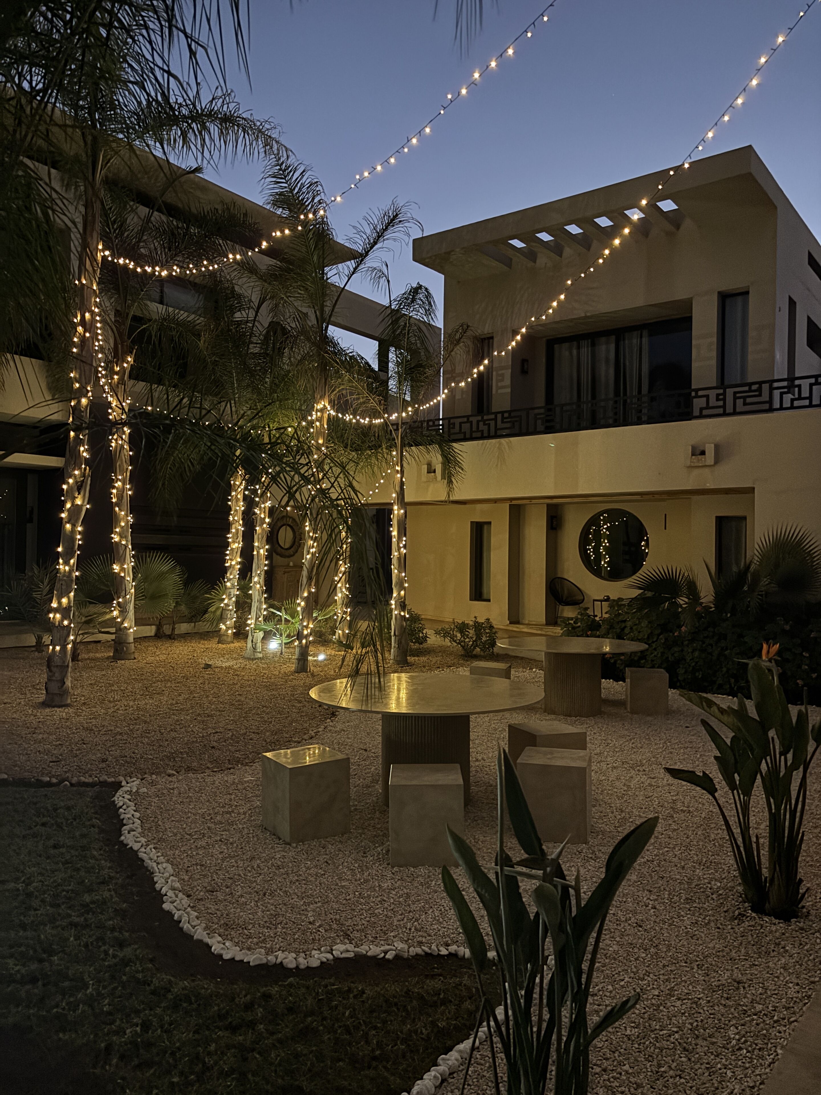 A courtyard with a round table and chairs, surrounded by palm trees and decorative lights.