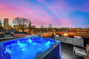 Una azotea con bañera de hidromasaje, mobiliario exterior y vistas a la ciudad al atardecer.