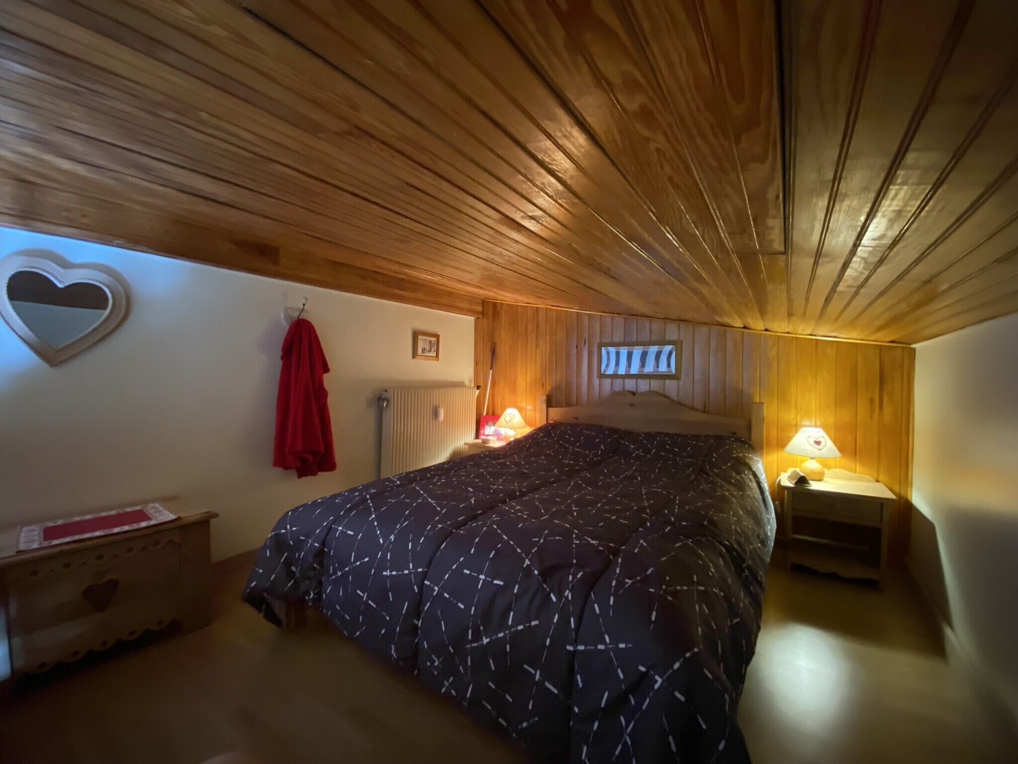 Un dormitorio acogedor con techo de madera, una cama con edredón estampado, una mesita de noche con lámpara y una decoración con forma de corazón en la pared.