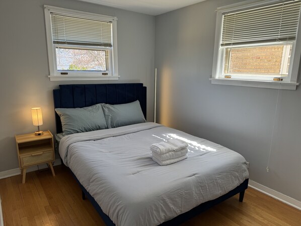 A bedroom with a bed, nightstand, lamp, and two windows with blinds.