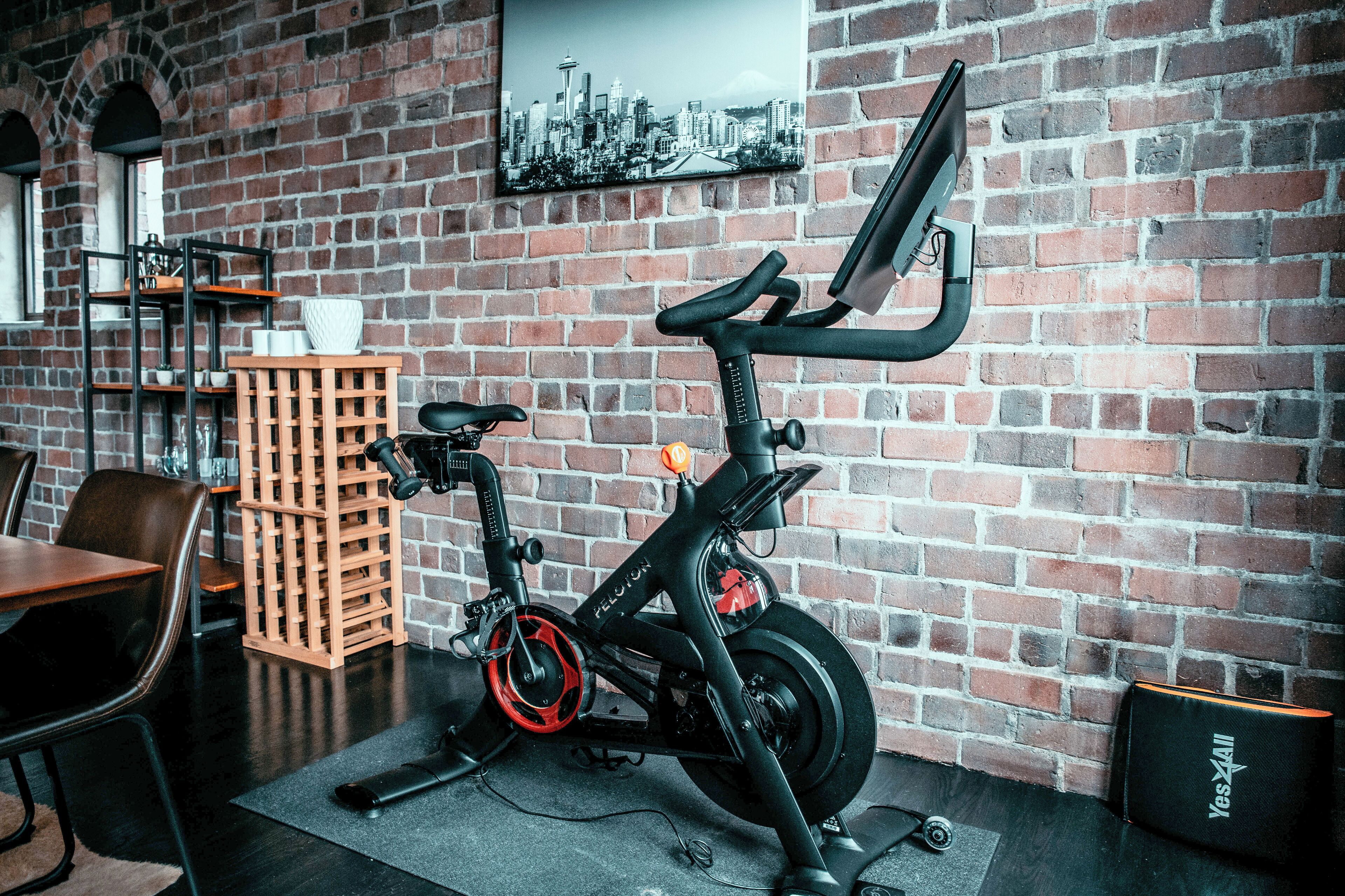 Una bicicleta estática en una habitación con paredes de ladrillo, un cuadro de un paisaje urbano y un caja de madera.